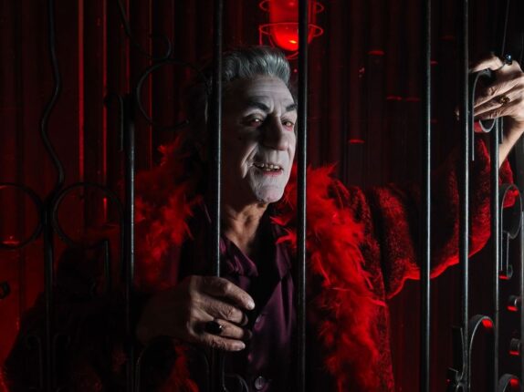 Gray-haired theatrical villain in red behind ornate iron bars