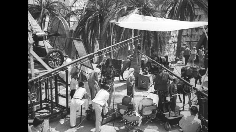 Black-and-white film set showing a camera crane, crew, and actors outdoors
